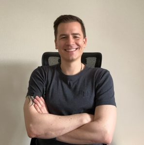 Der Autor Andreas Suchanek sitzt mit verschränken Armen vor der Kamera und lächelt.