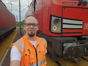 Auf dem Foto ist Lokführer Tim in einem hochsichtbaren orangefarbenen Eisenbahner-Warnweste. Er steht vor einer großen, roten Elektrolokomotive steht. Er lächelt und macht ein Selfie, mit der Kamera in seiner rechten Hand. Die Lokomotive gehört zur Deutschen Bahn (DB) und trägt das Logo von Siemens auf einer Trittstufe nahe dem Boden. Im Hintergrund sind weitere Teile des Zuges und Oberleitungen zu erkennen. Der Himmel ist bewölkt und es scheint ein Rangierbereich zu sein.