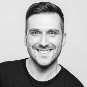 Das Portrait zeigt den Autor Manuel Merk. Das Foto wurde in einem Fotostudio aufgenommen. Manuel hat dunkle Haare und trägt ein schwarzes Oberteil. Er lächelt in die Kamera. Das Bild wurde von Fotografin Julia Klein aufgenommen.