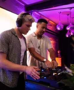 Auf dem Foto sind zwei Personen an einem DJ-Pult zu sehen. Eine der beiden trägt Kopfhörer und spielt Musik. Die andere Person mischt die Musik. Im Hintergrund ist ein Raum zu sehen der wie ein Club aussieht.