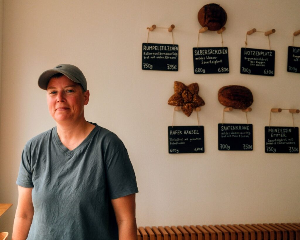 Auf dem Foto ist die autodidaktische Bäckerin Milena Drefke zu sehen. Sie steht am Fenstern in ihrem Café und hinter ihr sind einige Brote und Angebotstafeln zu sehen. Milena steht zum Fenster gewandt und trägt eine graue Kappe und ein graues T-Shirt. Sie lächelt in die Kamera. Das Foto wurde von Toni Borgert aufgenommen.