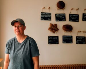 Auf dem Foto ist die autodidaktische Bäckerin Milena Drefke zu sehen. Sie steht am Fenstern in ihrem Café und hinter ihr sind einige Brote und Angebotstafeln zu sehen. Milena steht zum Fenster gewandt und trägt eine graue Kappe und ein graues T-Shirt. Sie lächelt in die Kamera. Das Foto wurde von Toni Borgert aufgenommen.