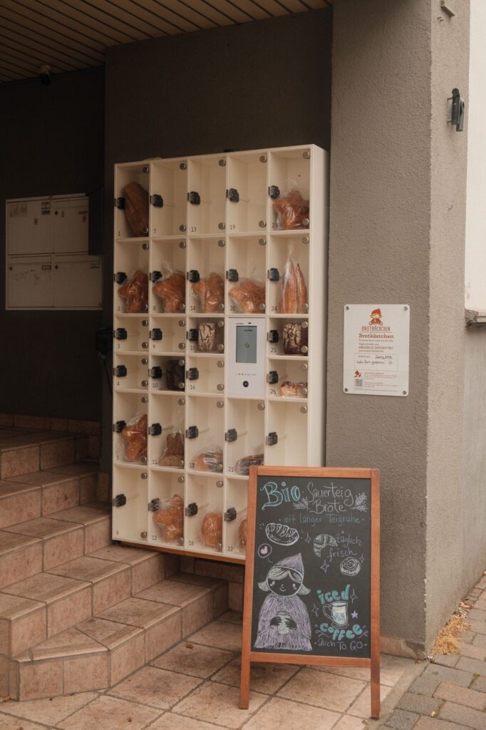 Das Foto zeigt eine sogenannte Markbox. Milena nutzt sie als Brotautomat. Es gibt 26 Fächer in die jeweils ein Brot mit Tüte passt. Die gesamte Box ist weiß, die Fächer haben eine transparente Tür. Genau in der Mitte ist ein Bezahlfeld für die Kartenzahlung zu sehen. Vor der Marktbox steht eine Kreidetafel mit der Werbung der Bäckerei. Die Box hängt über einer Treppe. Das Foto wurde von Toni Borgert aufgenommen.