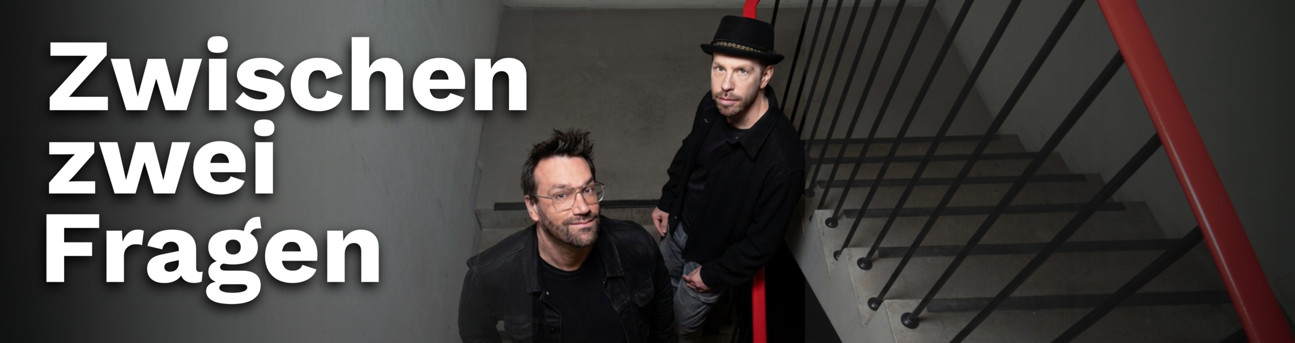 Das Foto zeigt das Cover vom Podcast "zwischen zwei Fragen" der von Toni und Sebastian gehostet wird. Die beiden stehen in einem Treppenhaus mit grauen Wänden und einem auffallend Roten Treppengeländer. Die beiden sind von oben fotografiert. Sebastian steht links, hat dunkle Haare, trägt eine Brille mit transparentem Rahmen und eine schwarze Jacke. Toni steht rechts etwas nach oben versetzt. Er trägt einen schwarzen Hut und eine schwarze Jacke sowie graue Hosen. Beide sehen mit neutralem Blick in die Kamera. Das Foto wurde von Tobias Jarmuzek aufgenommen.